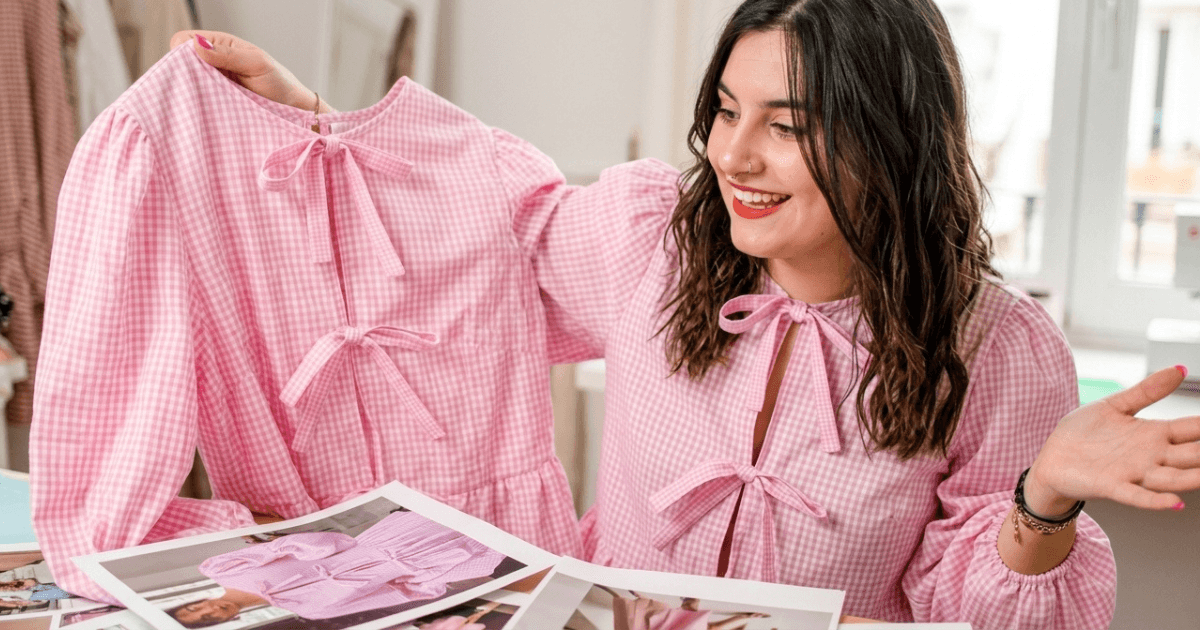 Mujer sonriente sostiene una prenda de vestir rosa con cuadros mientras muestra fotos de productos en una mesa, ilustrando la creación de imágenes de productos conIA.