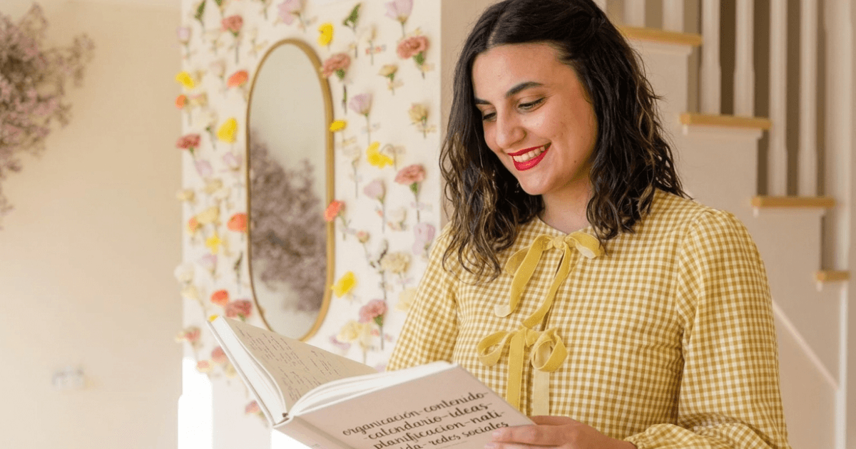 Mujer sonriente con blusa de cuadros amarillos sostiene un libro sobre organización de contenido, en un ambiente decorado con flores y un espejo ovalado.